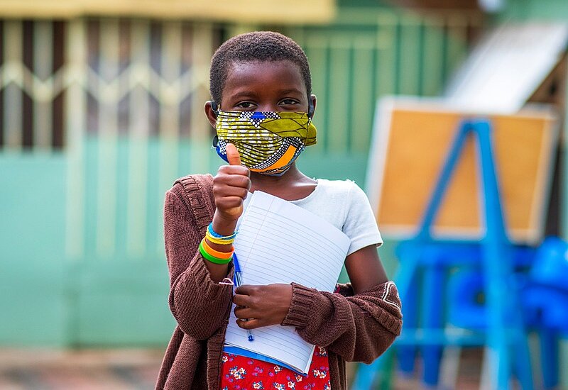Ein afrikanisches Mädchen mit einem bunten Tuch im Gesicht, hält im linken Arm ein Schreibblock und Stift und hält den rechten Daumen hoch.